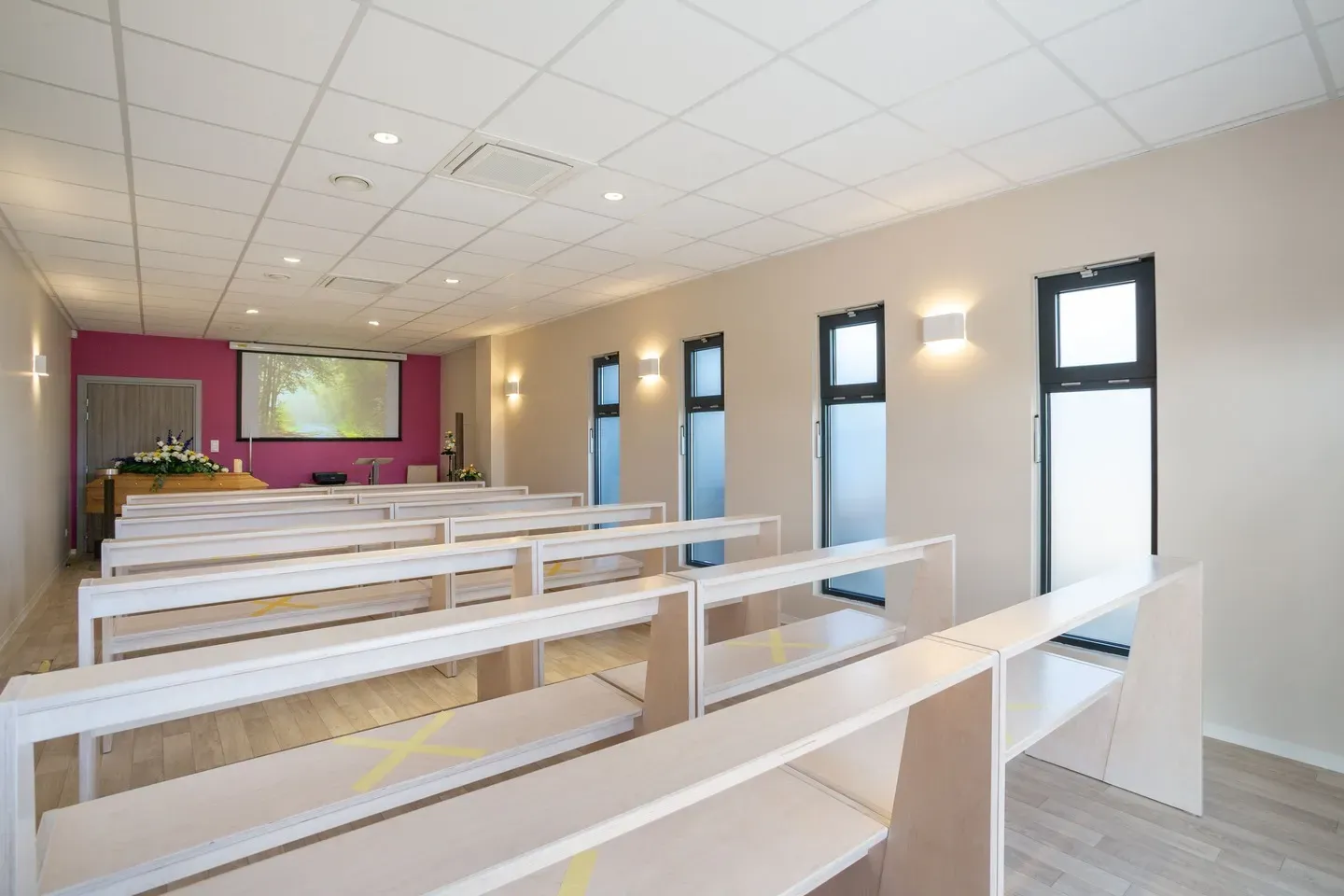 Salle de cérémonie funéraire moderne avec bancs blancs et écran de projection.